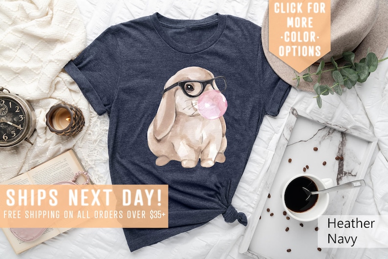 May include: A navy blue t-shirt with a watercolor illustration of a brown bunny wearing glasses and blowing a pink bubble. The text "SHIPS NEXT DAY! FREE SHIPPING ON ALL ORDERS OVER $35+" is at the bottom of the image.