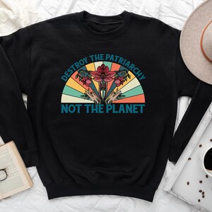 Op de afbeelding: Zwart sweatshirt met een vintage-stijl grafische afbeelding met de tekst "Destroy the Patriarchy Not the Planet". De afbeelding toont een bloem met handen die van onderaf omhoog reiken.