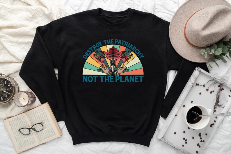 Op de afbeelding: Zwart sweatshirt met een vintage-stijl grafische afbeelding met de tekst "Destroy the Patriarchy Not the Planet". De afbeelding toont een bloem met handen die van onderaf omhoog reiken.