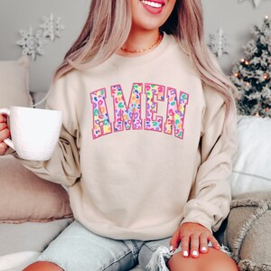 Puede incluir: Una sudadera beige con la palabra "AMEN" impresa en rosa con un dise&ntilde;o de estampado de leopardo arco&iacute;ris.