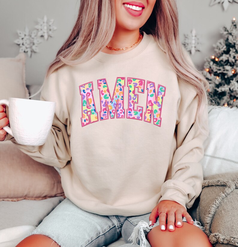Puede incluir: Una sudadera beige con la palabra "AMEN" impresa en rosa con un dise&ntilde;o de estampado de leopardo arco&iacute;ris.