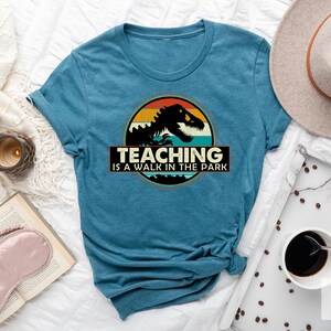May include: A teal blue t-shirt with a vintage-style graphic of a dinosaur in front of a sunset. The text "Teaching is a walk in the park" is printed below the graphic.