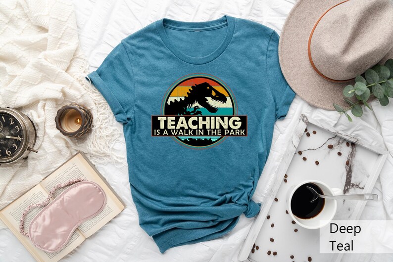 May include: A teal blue t-shirt with a vintage-style graphic of a dinosaur in front of a sunset. The text "Teaching is a walk in the park" is printed below the graphic.