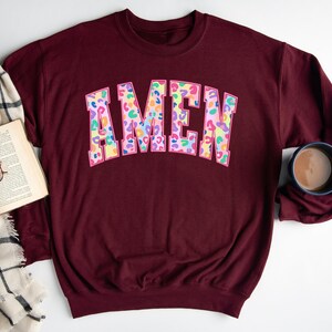 Puede incluir: Sudadera color burdeos con un gran gr&aacute;fico "AMEN" colorido en un dise&ntilde;o de estampado de leopardo.
