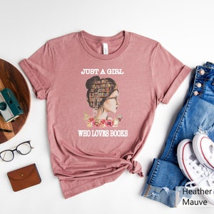 Pu&ograve; includere: Una t-shirt color malva heather con una grafica di una donna con una libreria come testa. Il testo "Just a girl who loves books" &egrave; stampato sulla t-shirt.