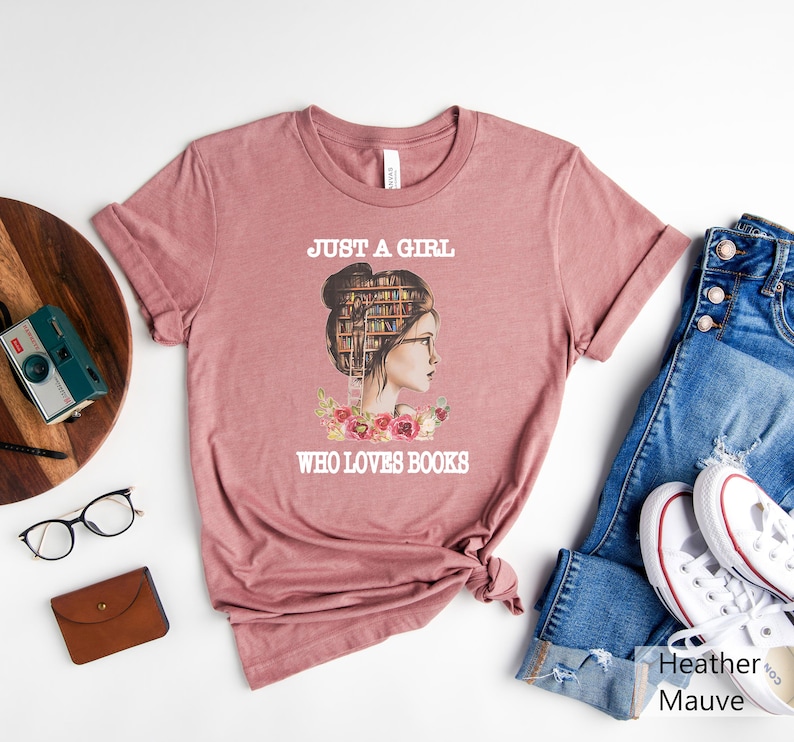 Pu&ograve; includere: Una t-shirt color malva heather con una grafica di una donna con una libreria come testa. Il testo "Just a girl who loves books" &egrave; stampato sulla t-shirt.