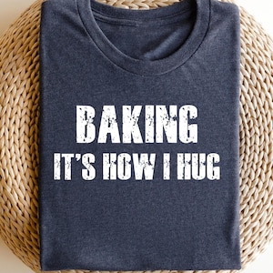May include: A navy blue t-shirt with the text "BAKING IT'S HOW I HUG" in distressed white lettering. The shirt is folded and resting on a woven, light brown surface.