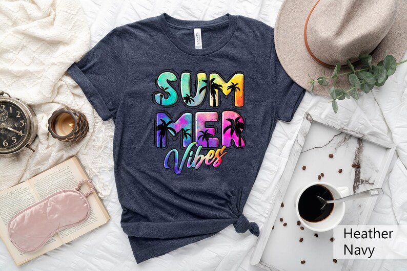 May include: Heather navy t-shirt with the words "SUMMER VIBES" in a colorful, tropical design. The design includes palm trees and a gradient of colors. A neutral-toned hat and a tray with coffee are also visible.