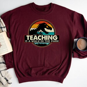 May include: Burgundy sweatshirt with a vintage-style graphic of a dinosaur with the text "Teaching is a walk in the park".