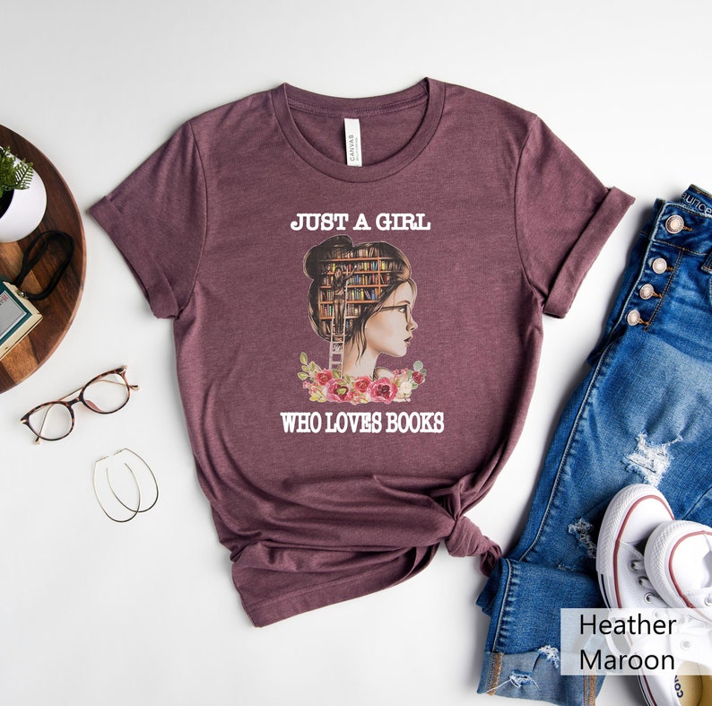 Pu&ograve; includere: Una t-shirt color marrone heather con una grafica di una donna con una libreria come testa. Il testo "Just a girl who loves books" &egrave; stampato sulla t-shirt.