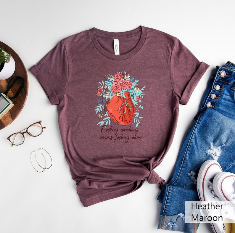 May include: A heather maroon t-shirt featuring a graphic of a heart surrounded by flowers. The text "Feeling emotions, means feeling alive" is printed below the heart.