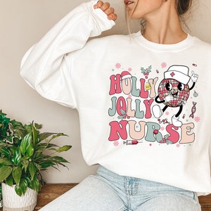 Op de afbeelding: Witte sweatshirt met de tekst "HOLLY JOLLY NURSE" in roze, rood en blauw retro lettertype. Een cartoon discobal karakter met een verpleegstersmuts is afgebeeld, samen met kerstthema-elementen.
