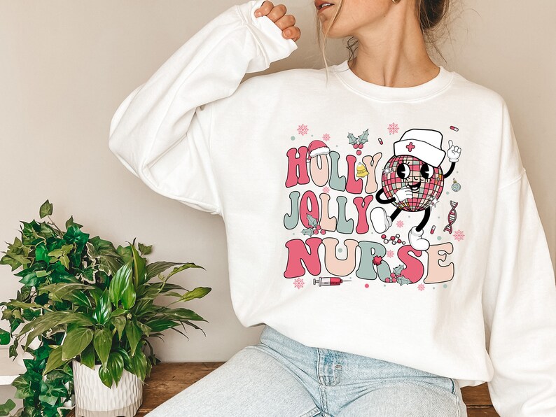 Op de afbeelding: Witte sweatshirt met de tekst "HOLLY JOLLY NURSE" in roze, rood en blauw retro lettertype. Een cartoon discobal karakter met een verpleegstersmuts is afgebeeld, samen met kerstthema-elementen.