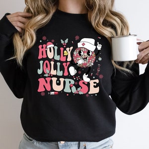 Op de afbeelding: Zwarte sweatshirt met de tekst "HOLLY JOLLY NURSE" in roze, teal en wit. Het ontwerp bevat een discobal-personage met een verpleegstershoed, hulst en sneeuwvlokken. Een witte mok wordt in de hand gehouden.