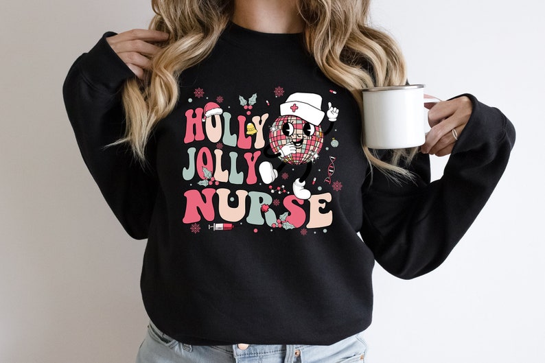 Op de afbeelding: Zwarte sweatshirt met de tekst "HOLLY JOLLY NURSE" in roze, teal en wit. Het ontwerp bevat een discobal-personage met een verpleegstershoed, hulst en sneeuwvlokken. Een witte mok wordt in de hand gehouden.