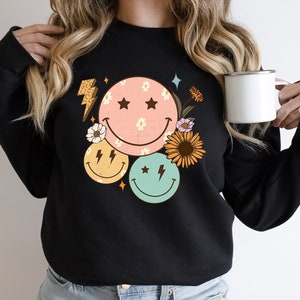 May include: Black sweatshirt with a graphic design of three colorful smiley faces with stars and lightning bolts, surrounded by flowers.