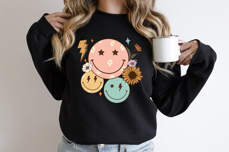 May include: Black sweatshirt with a graphic design of three colorful smiley faces with stars and lightning bolts, surrounded by flowers.