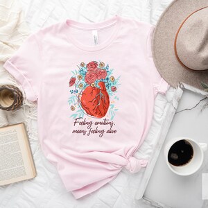 May include: Pink t-shirt featuring a graphic of a heart with flowers and the text "Feeling emotions, means feeling alive".