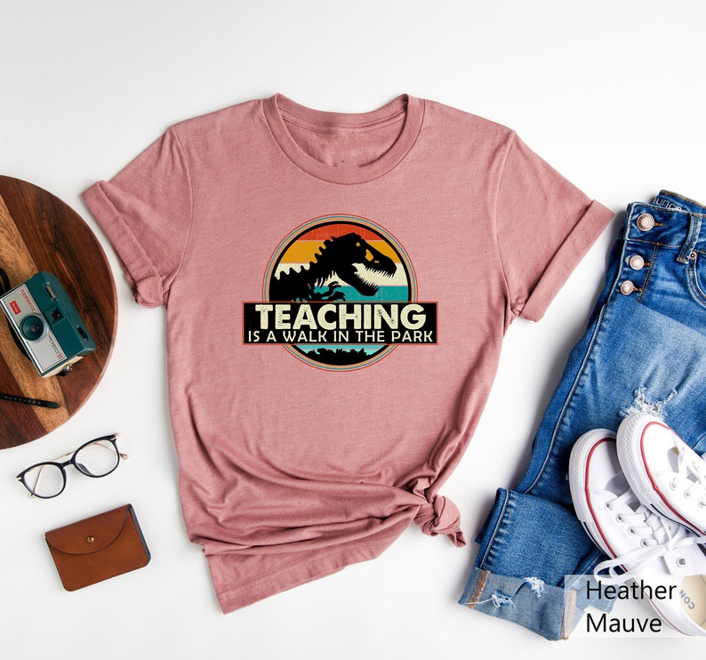 May include: A heather mauve t-shirt with a vintage-style graphic of a dinosaur with the text "Teaching is a walk in the park".