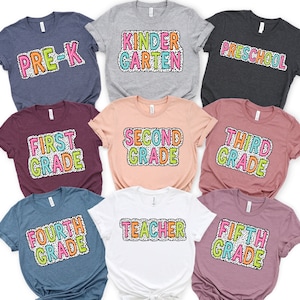 Puede incluir: Nueve camisetas diferentes con texto colorido sobre un fondo blanco con lunares negros. Las camisetas están etiquetadas con diferentes niveles de grado: Pre-K, Kindergarten, Preschool, First Grade, Second Grade, Third Grade, Fourth Grade, Fifth Grade y Teacher.
