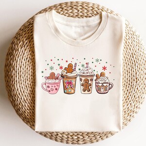 Puede incluir: Una camiseta blanca con un dise&ntilde;o festivo que presenta cuatro hombres de jengibre sosteniendo tazas de chocolate caliente cubiertas con crema batida y chispas. Las tazas est&aacute;n decoradas con bastones de caramelo y casas de jengibre.