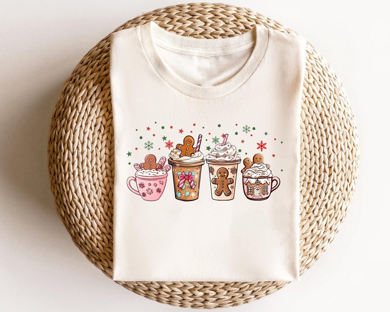 Puede incluir: Una camiseta blanca con un dise&ntilde;o festivo que presenta cuatro hombres de jengibre sosteniendo tazas de chocolate caliente cubiertas con crema batida y chispas. Las tazas est&aacute;n decoradas con bastones de caramelo y casas de jengibre.