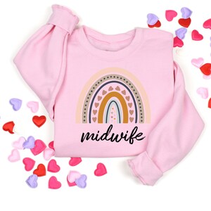 Peut inclure: Sweat-shirt rose avec un motif arc-en-ciel et le mot "sage-femme" imprim&eacute; sur le devant. Le sweat-shirt est entour&eacute; de petits c&oelig;urs roses, rouges et violets.