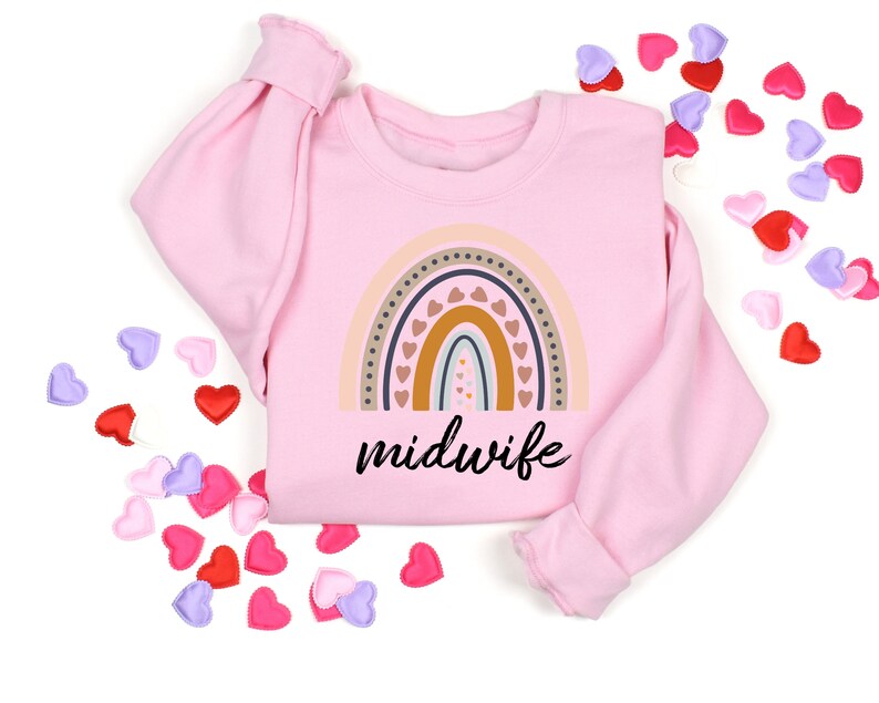 Peut inclure: Sweat-shirt rose avec un motif arc-en-ciel et le mot "sage-femme" imprim&eacute; sur le devant. Le sweat-shirt est entour&eacute; de petits c&oelig;urs roses, rouges et violets.