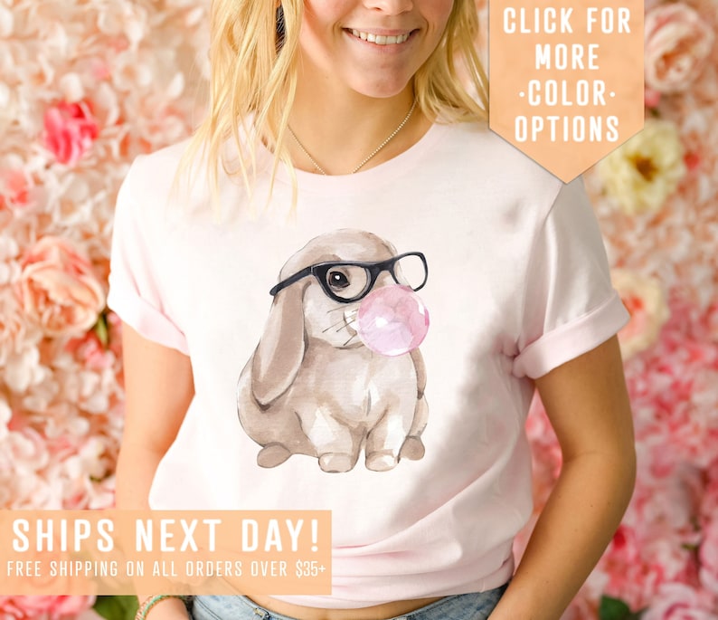 May include: Pink t-shirt with a watercolor illustration of a brown bunny wearing black glasses and blowing a pink bubble. The text "SHIPS NEXT DAY! FREE SHIPPING ON ALL ORDERS OVER $35+" is at the bottom of the shirt. The text "CLICK FOR MORE -COLOR- OPTIONS" is in the top right corner of the image.