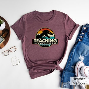 May include: Heather maroon t-shirt with a vintage-style graphic of a dinosaur in front of a sunset. The text "Teaching is a walk in the park" is printed below the graphic.