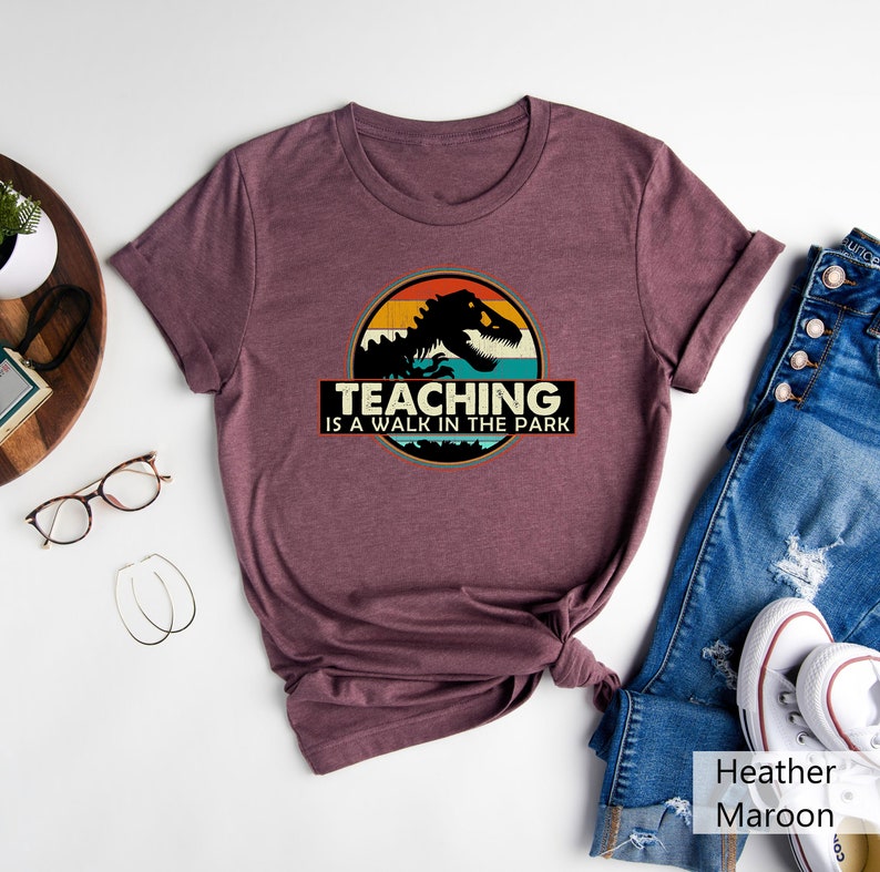 May include: Heather maroon t-shirt with a vintage-style graphic of a dinosaur in front of a sunset. The text "Teaching is a walk in the park" is printed below the graphic.