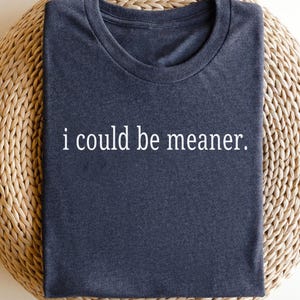 May include: Dark blue t-shirt with the phrase "i could be meaner" in white, centered on the chest. The shirt is folded and resting on a woven, light brown surface.