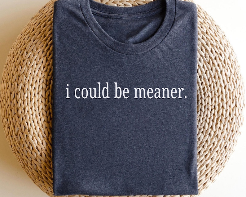 May include: Dark blue t-shirt with the phrase "i could be meaner" in white, centered on the chest. The shirt is folded and resting on a woven, light brown surface.