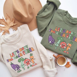 May include: Two cream and olive green sweatshirts with the text "Let's root for each other" and a graphic of cartoon vegetables.