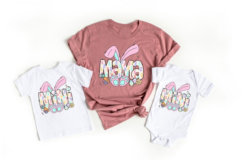 Pu&ograve; includere: Un set di tre magliette con un design di coniglio pasquale. Le magliette dicono "Mama" e "Mini" in un font colorato e a motivi. Le magliette sono bianche con orecchie di coniglio rosa e piedi di coniglio blu.