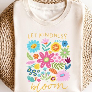 May include: A white t-shirt with the text "Let Kindness Bloom" printed in yellow. The text is surrounded by a colorful floral design with pink, yellow, blue, green, and orange flowers.