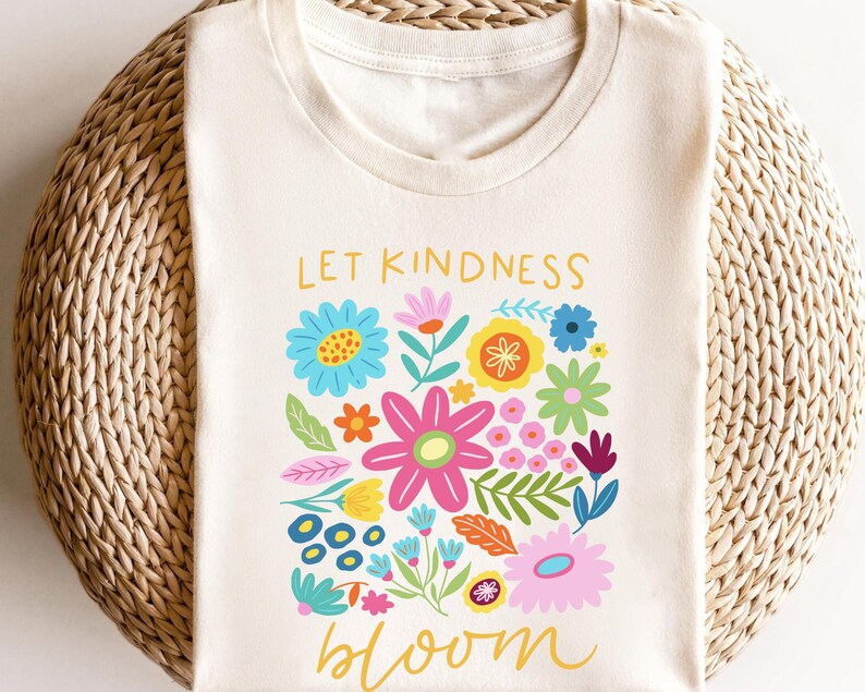 May include: A white t-shirt with the text "Let Kindness Bloom" printed in yellow. The text is surrounded by a colorful floral design with pink, yellow, blue, green, and orange flowers.