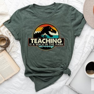 May include: A green t-shirt with a vintage-style graphic of a dinosaur in a sunset. The text "TEACHING IS A WALK IN THE PARK" is printed below the graphic.