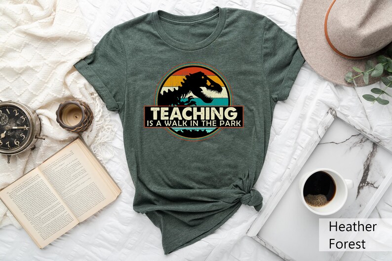 May include: A green t-shirt with a vintage-style graphic of a dinosaur in a sunset. The text "TEACHING IS A WALK IN THE PARK" is printed below the graphic.