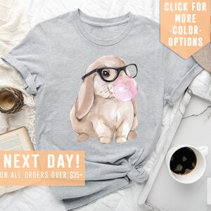 May include: Gray t-shirt with a watercolor illustration of a brown bunny wearing glasses and blowing a pink bubble. The text "SHIPS NEXT DAY! FREE SHIPPING ON ALL ORDERS OVER $35+" is at the bottom of the image.