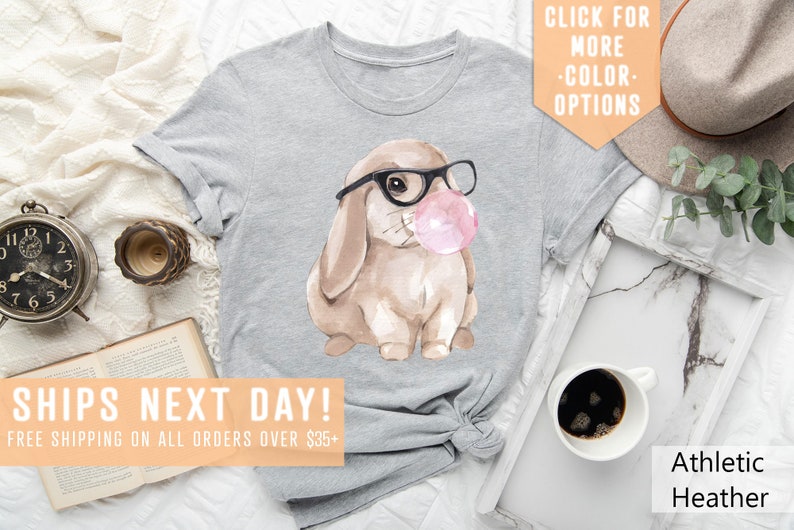 May include: Gray t-shirt with a watercolor illustration of a brown bunny wearing glasses and blowing a pink bubble. The text "SHIPS NEXT DAY! FREE SHIPPING ON ALL ORDERS OVER $35+" is at the bottom of the image.