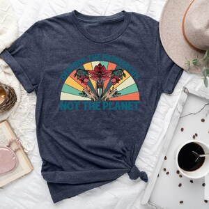 Op de afbeelding: Een marineblauw t-shirt met een retro-stijl grafische afbeelding met de tekst "Vernietig het patriarchaat, niet de planeet". De afbeelding toont een gestileerde baarmoeder met bloemen en handen die omhoog reiken.