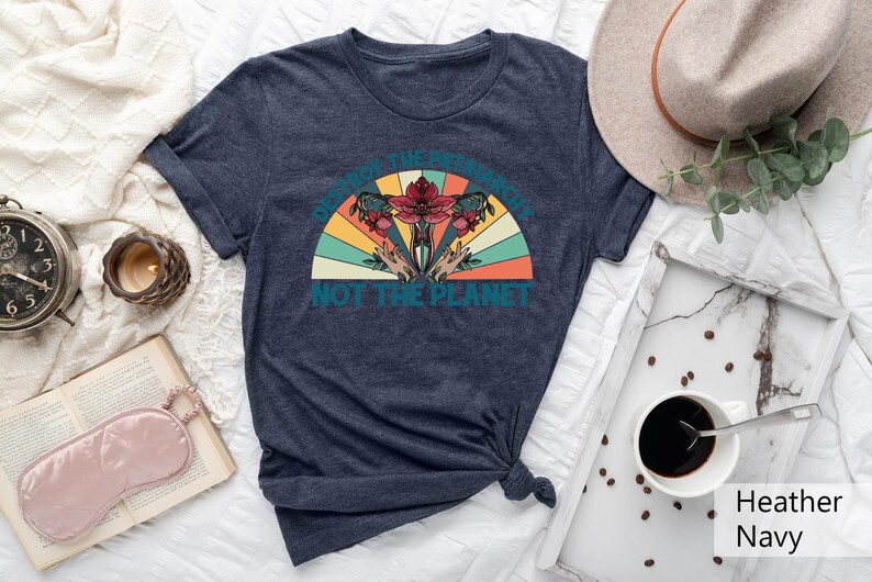 Op de afbeelding: Een marineblauw t-shirt met een retro-stijl grafische afbeelding met de tekst "Vernietig het patriarchaat, niet de planeet". De afbeelding toont een gestileerde baarmoeder met bloemen en handen die omhoog reiken.