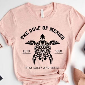 May include: A light pink t-shirt with a black graphic of a sea turtle and the text "THE GULF OF MEXICO" above it. Below the turtle is the text "ESTD 1550" and "STAY SALTY AND RESIST".