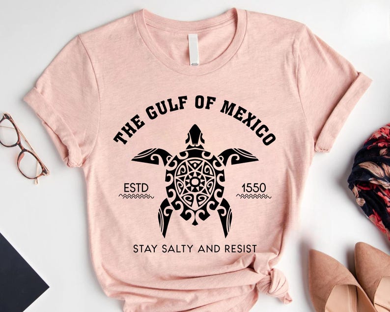 May include: A light pink t-shirt with a black graphic of a sea turtle and the text "THE GULF OF MEXICO" above it. Below the turtle is the text "ESTD 1550" and "STAY SALTY AND RESIST".