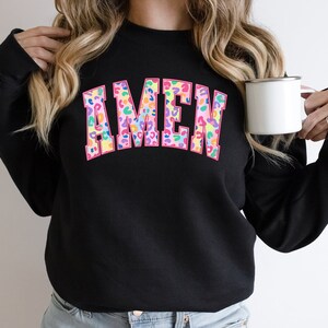 Puede incluir: Sudadera negra con la palabra "AMEN" en letras grandes de color rosa con un estampado de leopardo colorido.