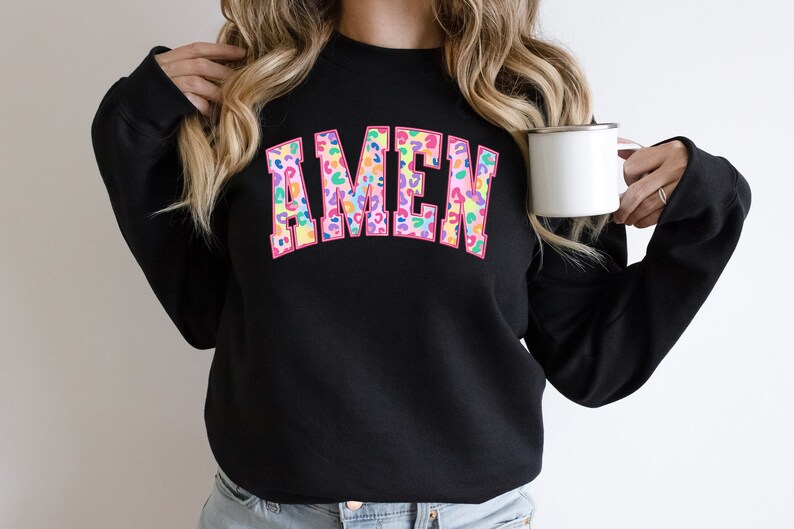 Puede incluir: Sudadera negra con la palabra "AMEN" en letras grandes de color rosa con un estampado de leopardo colorido.