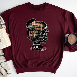 May include: A maroon sweatshirt with a graphic design of a woman's silhouette with a bookshelf behind her. The text reads "& Into the library she goes to lose her MIND and find her SOUL."
