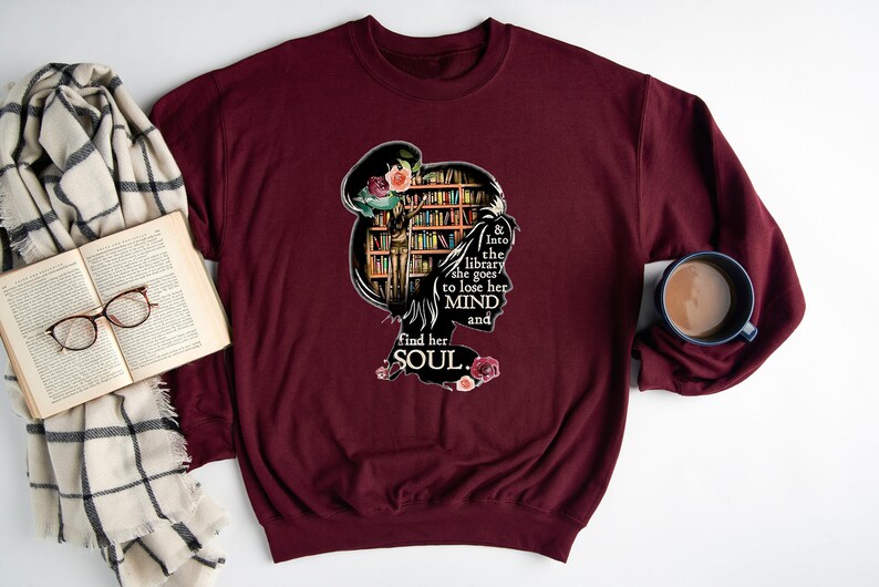 May include: A maroon sweatshirt with a graphic design of a woman's silhouette with a bookshelf behind her. The text reads "& Into the library she goes to lose her MIND and find her SOUL."