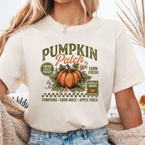 May include: A cream-colored short-sleeve t-shirt featuring a graphic of an orange pumpkin surrounded by green leaves. The text reads PUMPKIN Patch, FRESH FROM THE PATCH, FARM FRESH, LOCALLY GROWN, AUTUMN PICKED, PUMPKINS, CORN MAZE, APPLE CIDER.